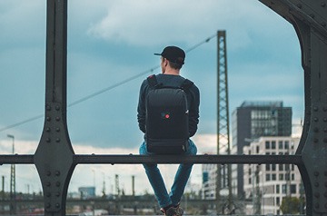Man with a backpack sitting on a bridge looking away from us toward a cityscape