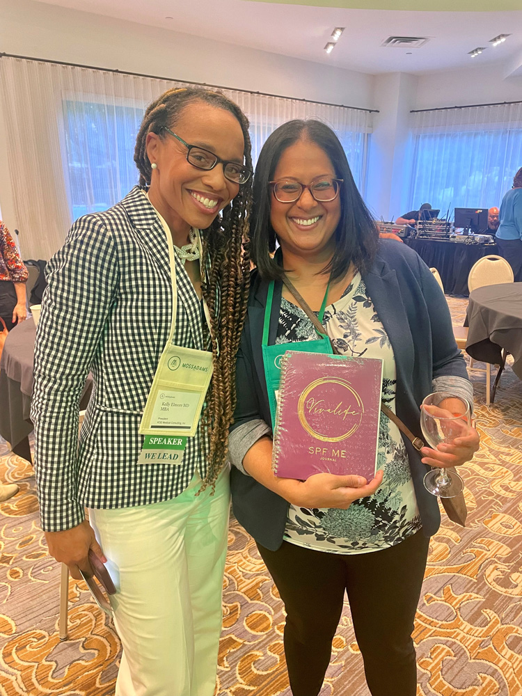 Dr Kelly Elmore (Panelist) and Aparna Venkateswaran: Two women gather for a photo in front of a conference room of other attendees.