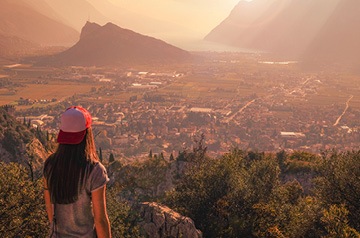 Woman in a ballcap standing on a hill overlooking a small city surrounded by mountains beside the ocean.