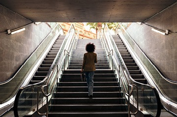 Seen from the back, a woman walking up a staircase that’s between two escalators.