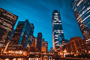 A view of skyscrapers with their lights on against a dark blue evening sky.