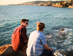 two men sitting on coastal rocks, waves below