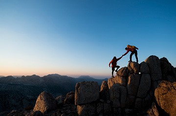 One hiker helping a second hiker ascend the top of a rock peak