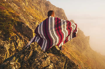 person wrapped in knit blanket sitting on rocky cliff