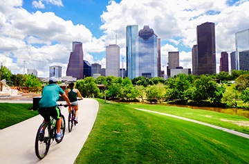 Two people riding bicycles on a path in a park with a cityscape in the background